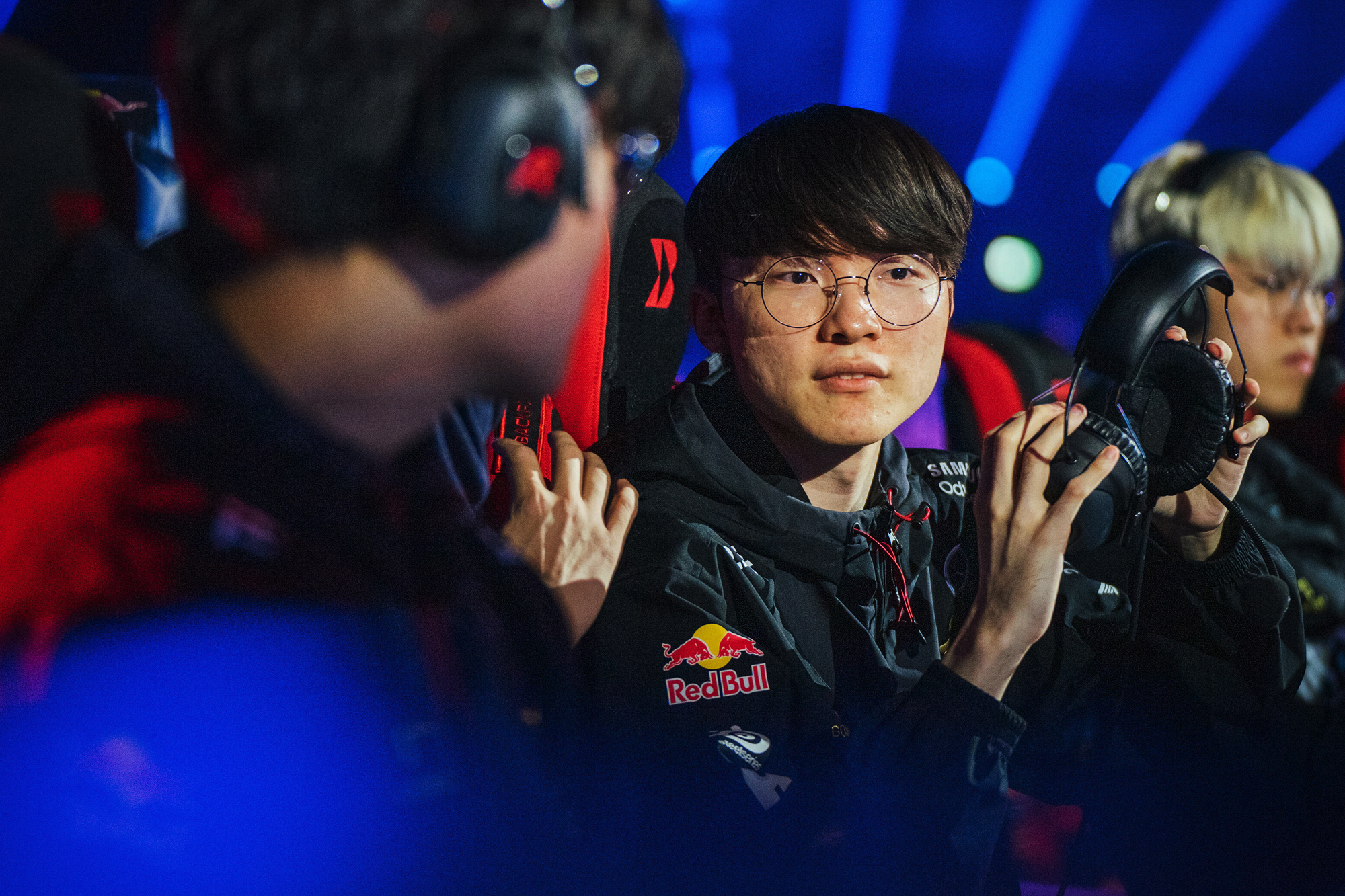 A young esports player wearing glasses and a headset sits in front of a computer, holding his headphones, while another player in a Red Bull LOIO jacket speaks to him. Blue stage lights glow in the background.
