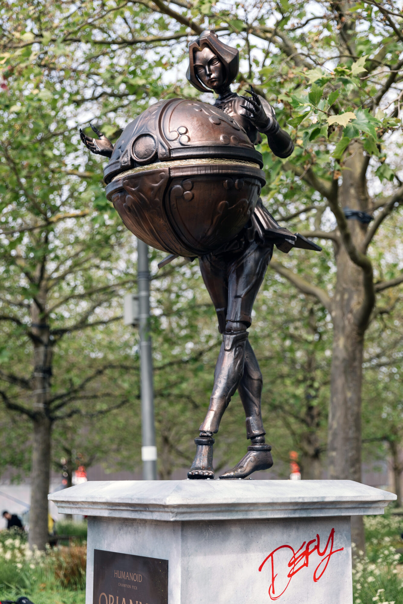 A bronze statue of a futuristic, humanoid figure with a large spherical object around its torso stands on a pedestal outdoors in a park, reminiscent of the iconic trophies awarded at Riot Games MSI events.
