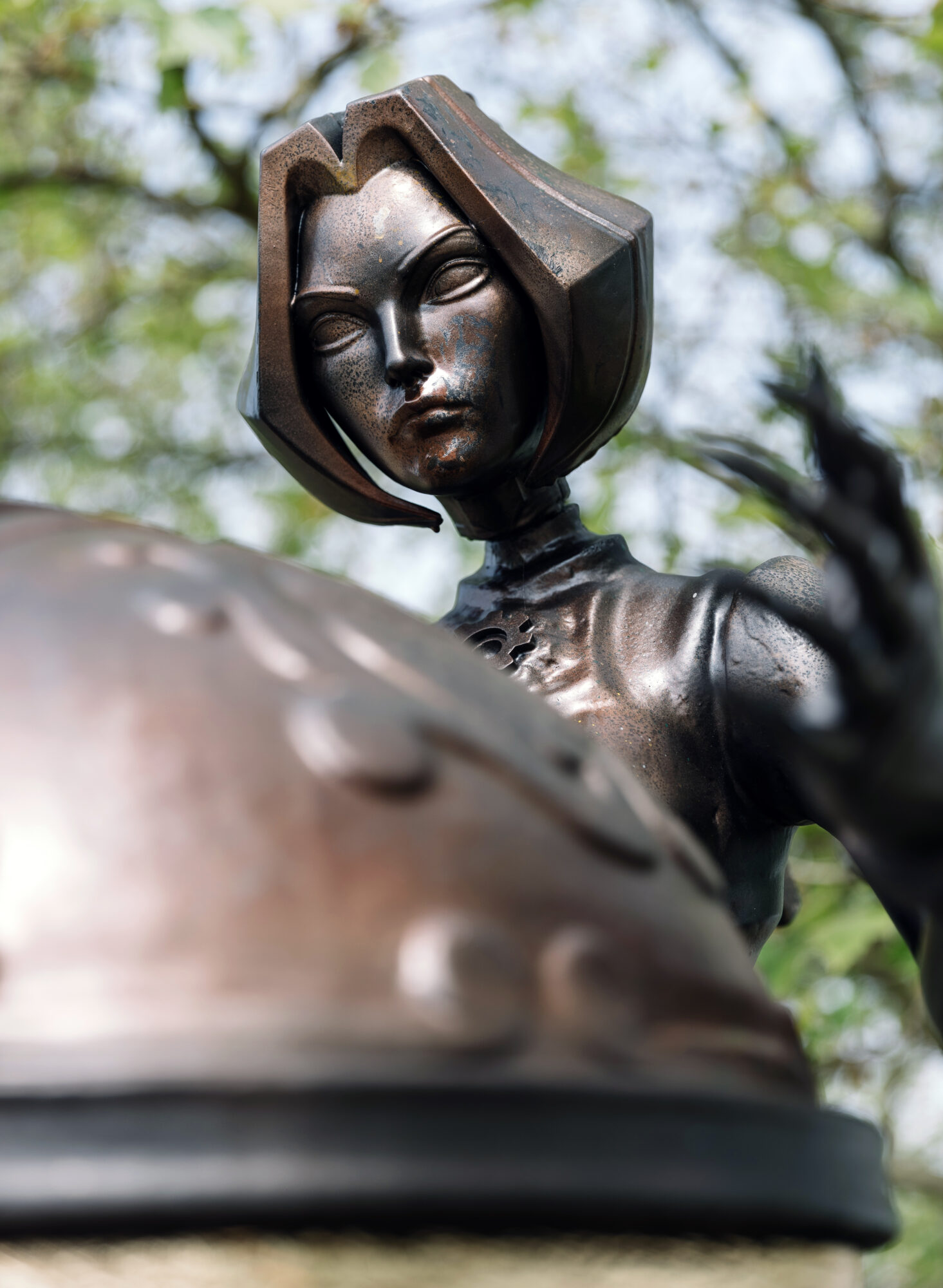 A close-up view of a metallic statue of a woman with a stylized, angular face and short hair, looking down thoughtfully with her hand raised—reminiscent of the artistry seen at Riot Games MSI—set against blurred green foliage.