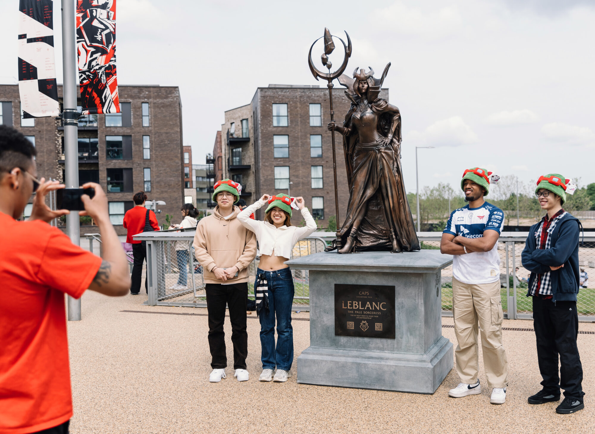Four people wearing festive hats pose for a photo next to a large statue of LeBlanc from League of Legends in an urban outdoor area during the Riot Games MSI, while another person takes their picture.