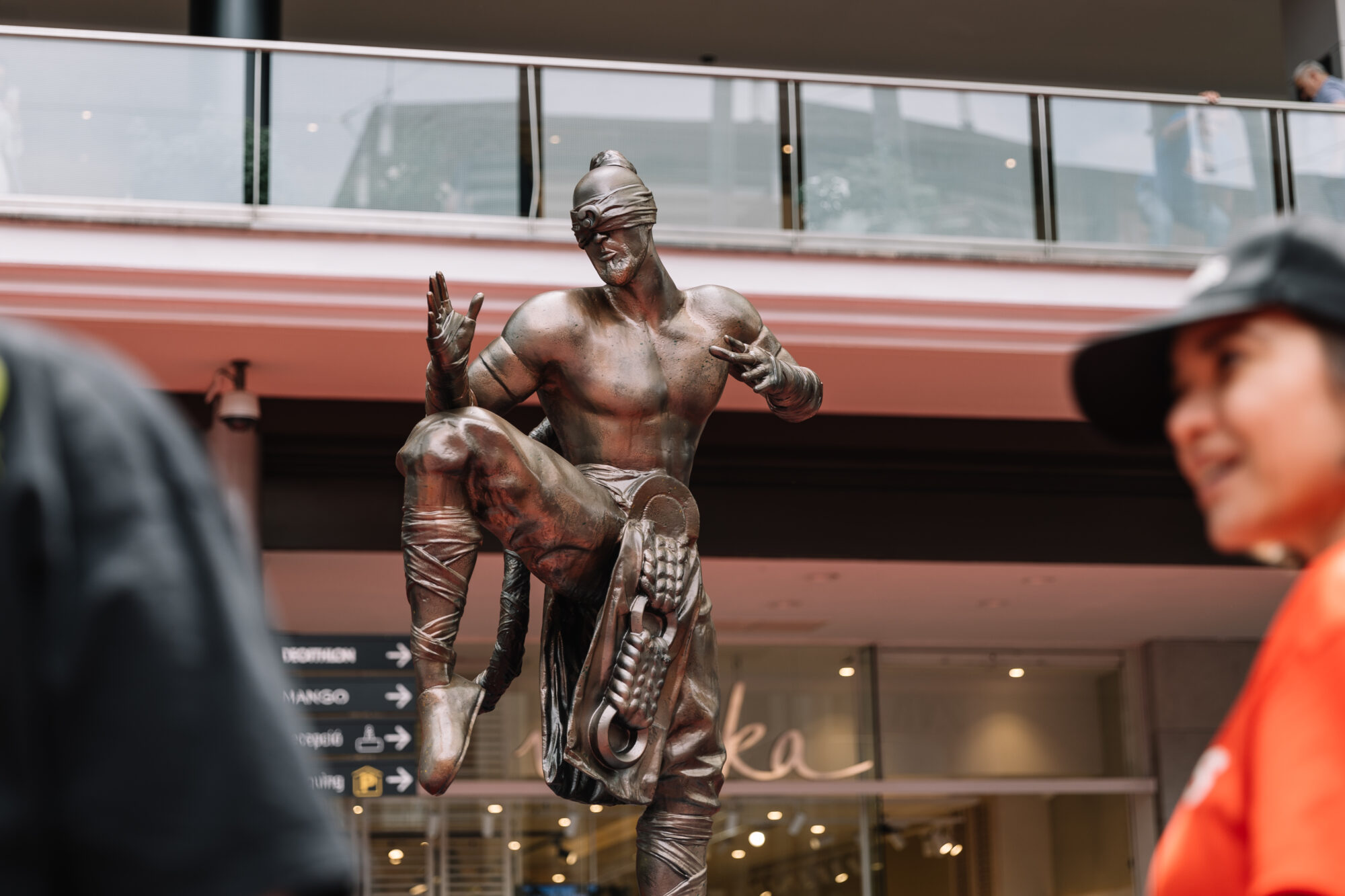 A bronze street performer dressed as a statue poses with one leg raised and fingers pointing, wearing a headband and rollerblades, while people pass by in the foreground at the lively Riot Games MSI event.