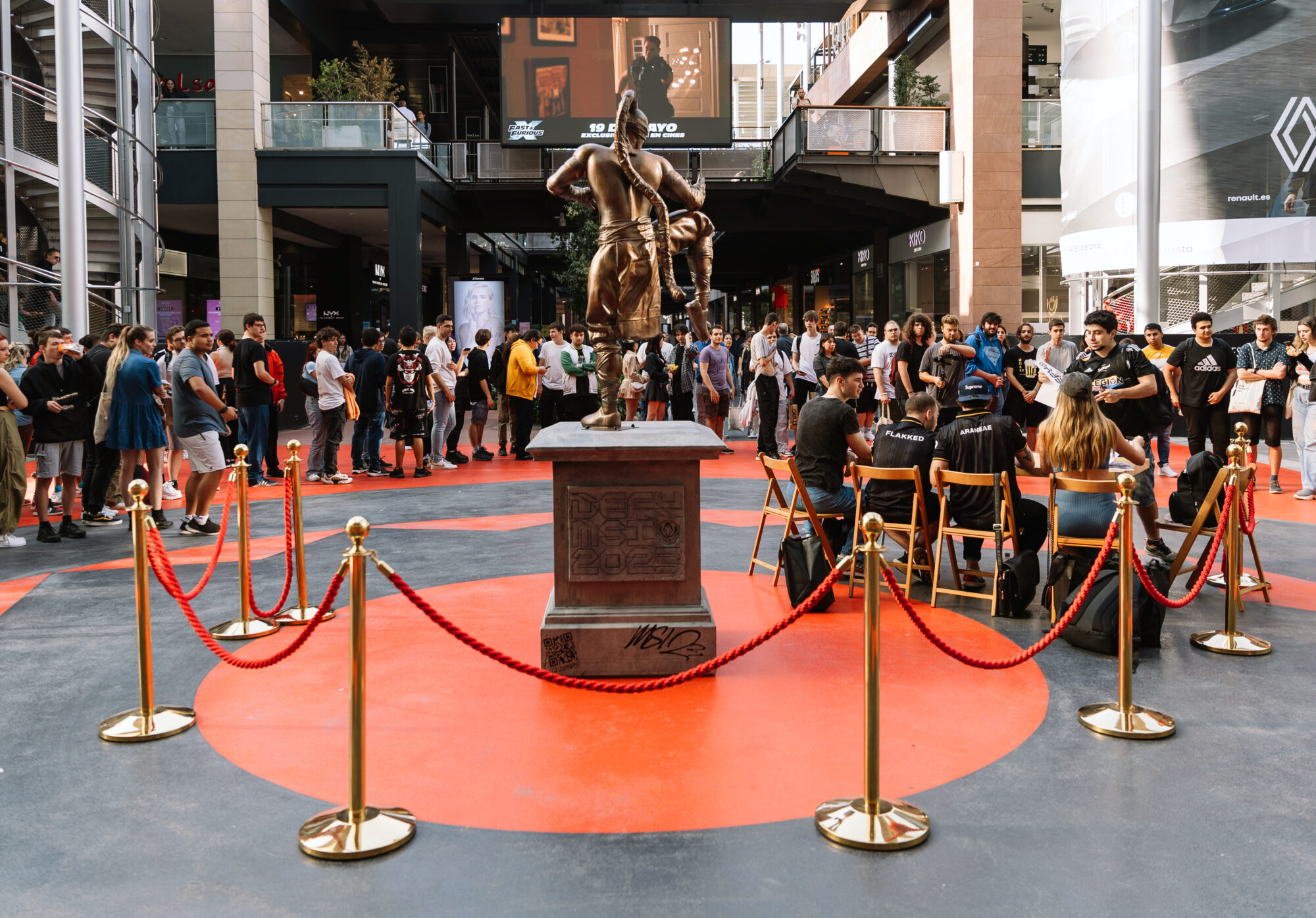 A crowd gathers around a statue enclosed by red ropes in an urban plaza during the Riot Games MSI event. Several people sit on chairs near the statue, while others stand and watch. Modern buildings and large screens are in the background.