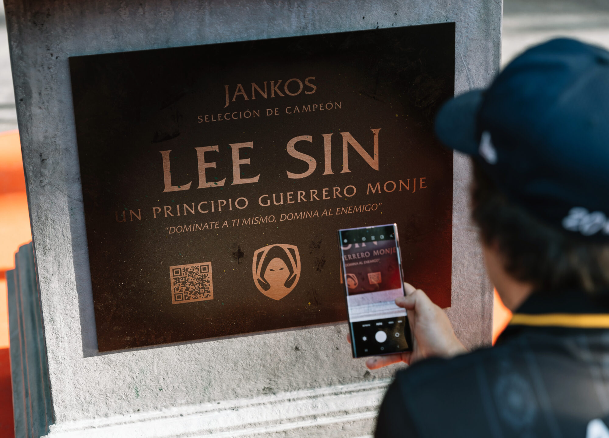 A person holding a smartphone takes a photo of a plaque dedicated to Lee Sin, labeled as a warrior monk, featuring text in Spanish, an engraved face, a QR code on the plaque, and a nod to Riot Games MSI.