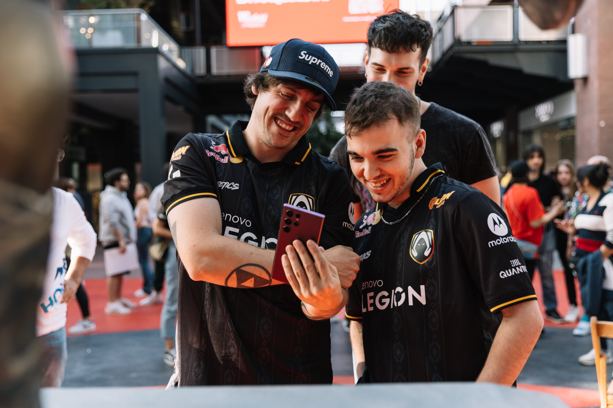 Three young men wearing matching black esports jerseys smile and laugh as they look at a phone together outdoors during Riot Games MSI, surrounded by a crowd of people in a lively, urban setting.