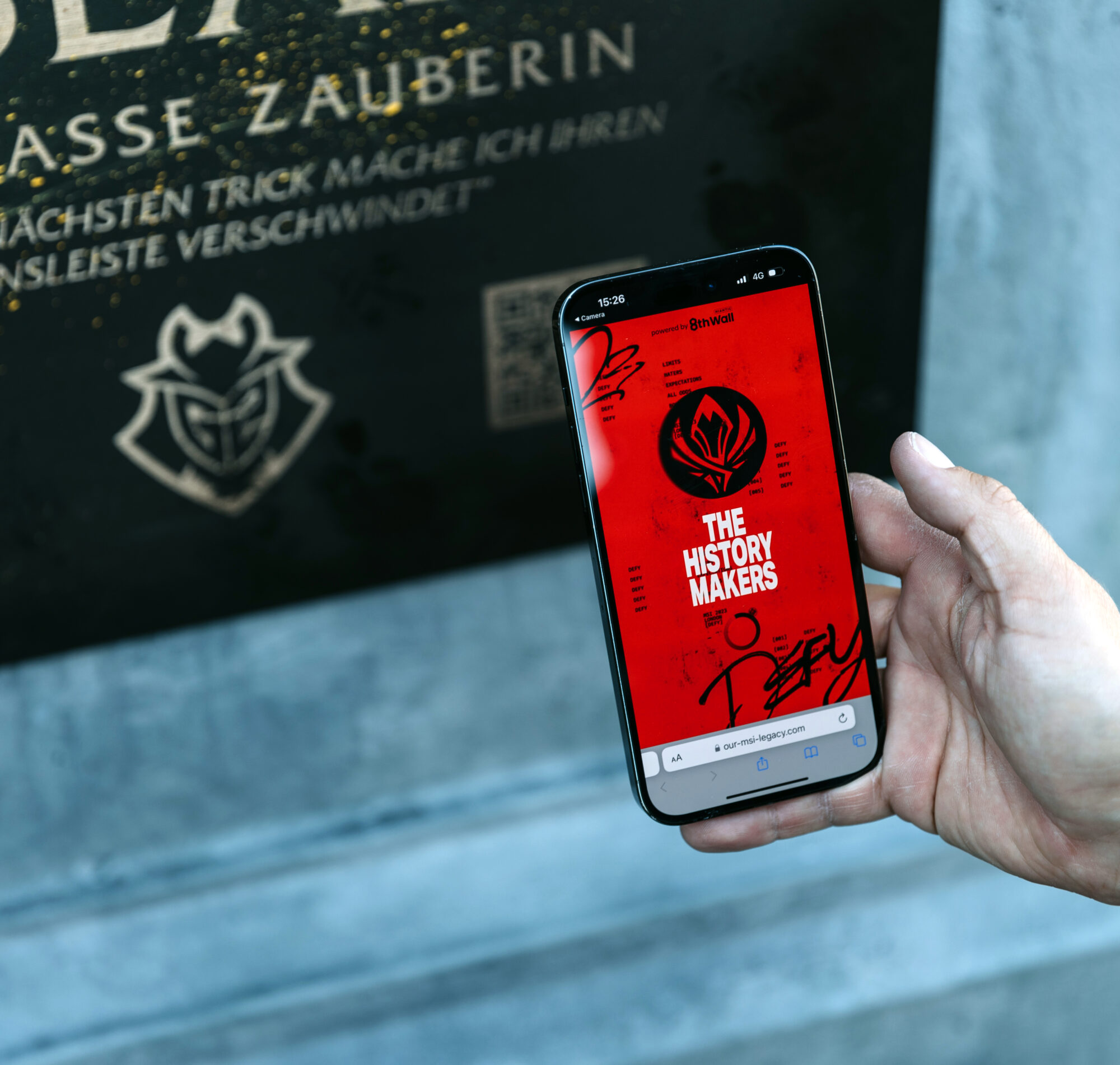 A hand holds a smartphone displaying a red screen with the text THE HISTORY MAKERS beneath a stylized logo, next to a black and gold plaque with partially visible German text—capturing the spirit of Riot Games MSI moments.