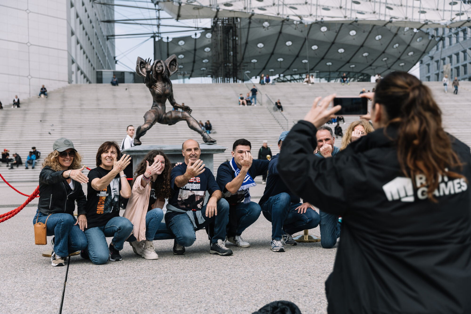 A group of people posing and smiling for a photo in front of a large statue and modern building, while a woman in a jacket with “Rallye Games” on the back captures the moment at Riot Games MSI.