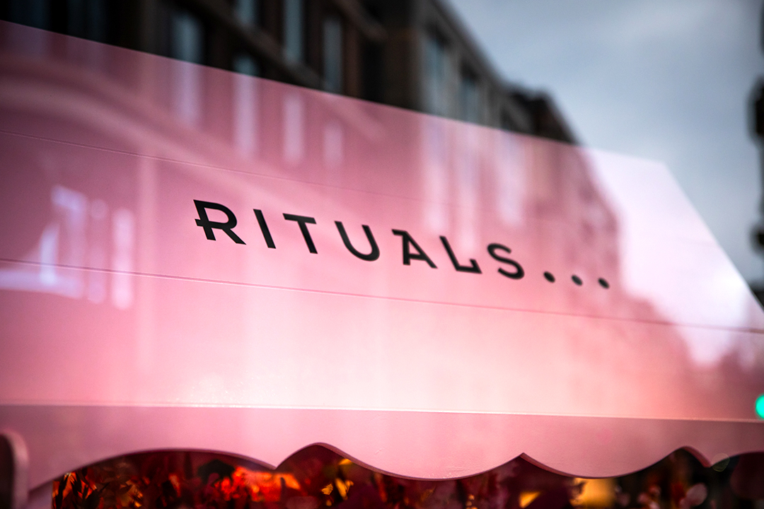 A close-up of a pink awning with the words RITUALS... printed in bold black letters, highlighting a special Rituals Mother’s Day display, with buildings blurred in the background.