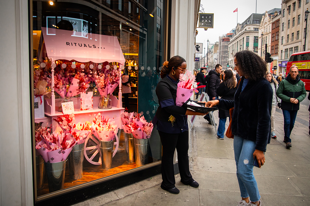 A woman hands a bouquet of pink flowers to another woman outside a Rituals store decorated with pink blooms for Rituals Mother’s Day, while pedestrians stroll past on the bustling city street.