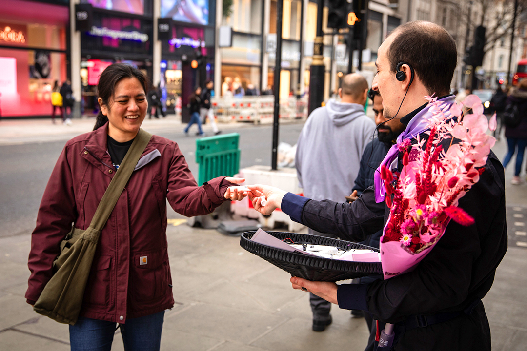 A smiling woman in a maroon jacket accepts a small item from a man holding a bouquet of pink flowers and a tray on a busy city street, capturing the joyful rituals of Mother’s Day amid bustling pedestrians and storefronts.