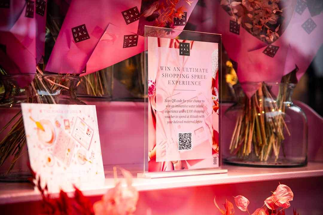 A display with pink flower bouquets in glass vases and a sign promoting a Rituals Mother’s Day shopping spree contest. The sign features a QR code, surrounded by soft pink decorations and floral arrangements.