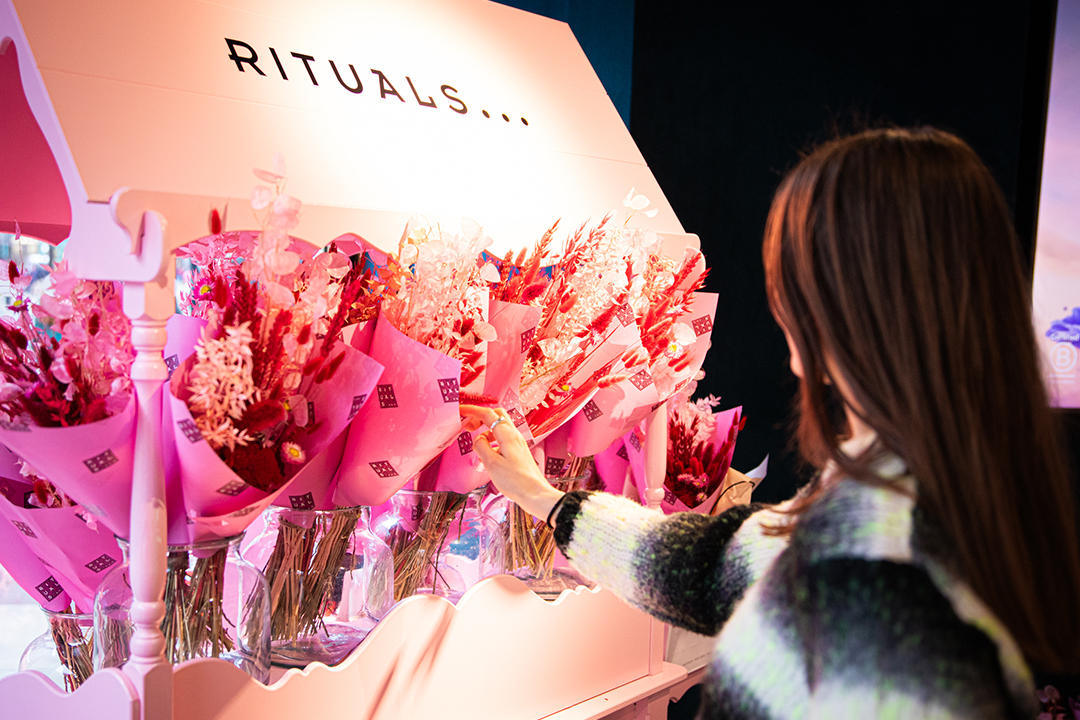 A woman with brown hair selects a bouquet of pink flowers from a display under a sign that reads RITUALS in a well-lit store, celebrating Rituals Mother’s Day.