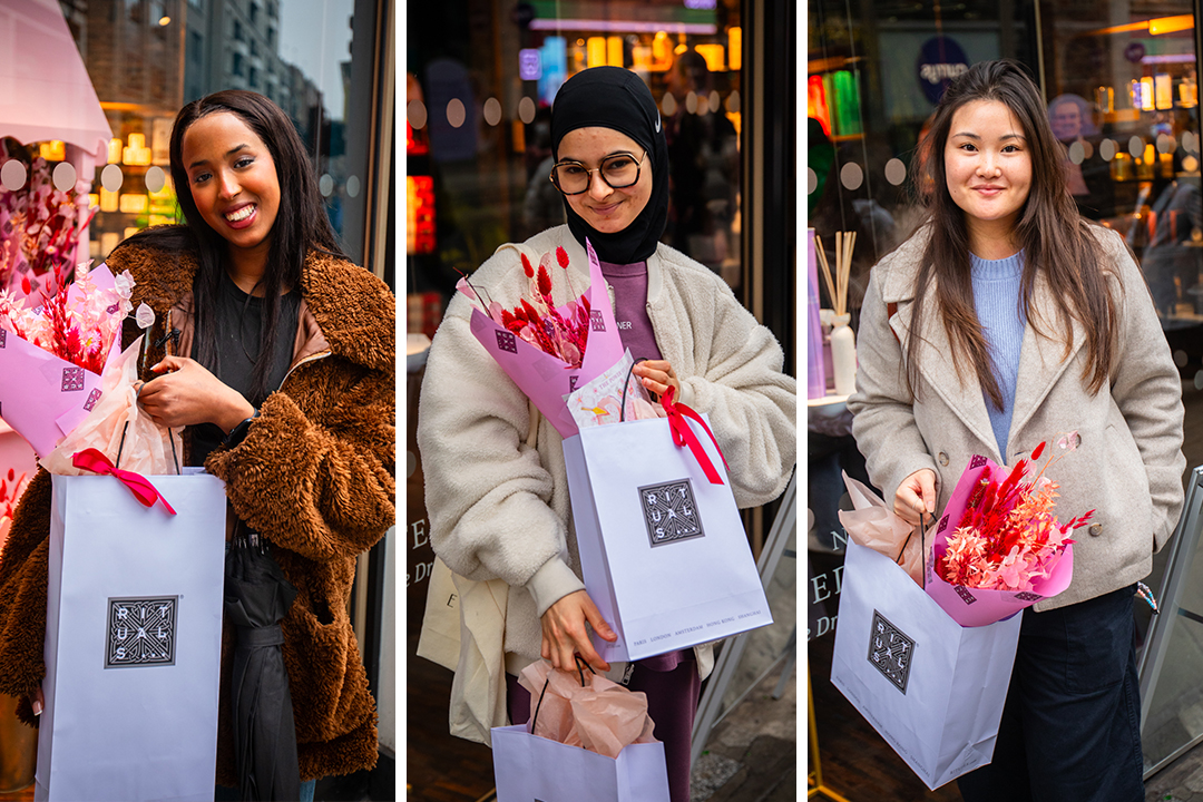 Three women stand outside a store, each holding a white Rituals shopping bag and a bouquet of pink flowers, smiling for the camera. Urban glass windows and colorful lights set the scene for their joyful Rituals Mother’s Day celebration.