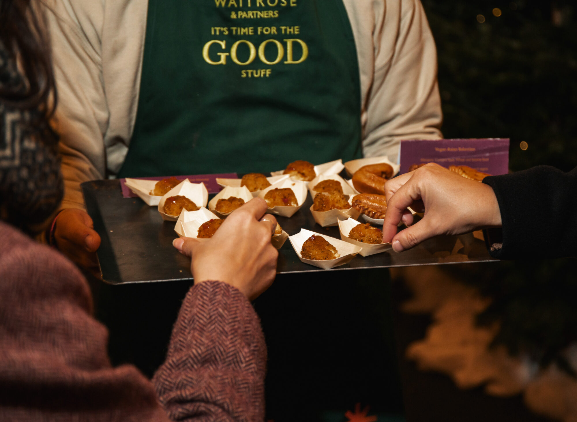 A person wearing a green apron holds a tray of small food samples in paper cups, while others reach to take them at the Waitrose Winter Festival tasting event.