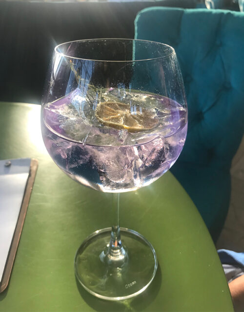 A large wine glass filled with a light purple drink and ice cubes sits on a green table, basking in sunlight—the perfect refreshment for a marketing team brainstorming session, with a teal upholstered chair in the background.