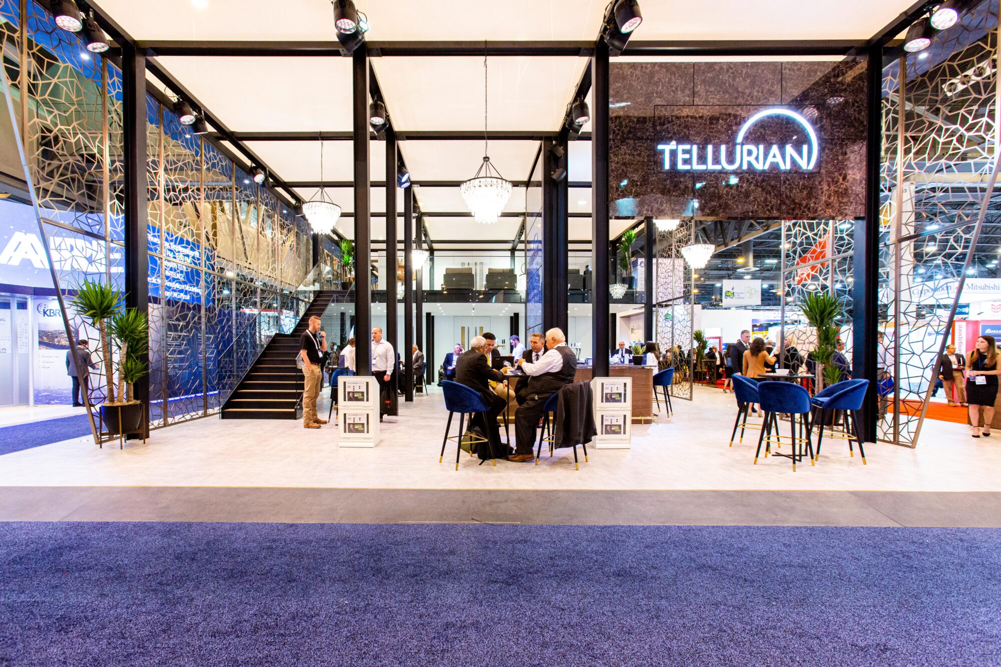 Modern exhibition booth with geometric glass walls, blue chairs, and people at tables. The illuminated TELLURIAN logo stands out above, creating a dynamic scene worthy of a Tellurian case study at this bustling expo.