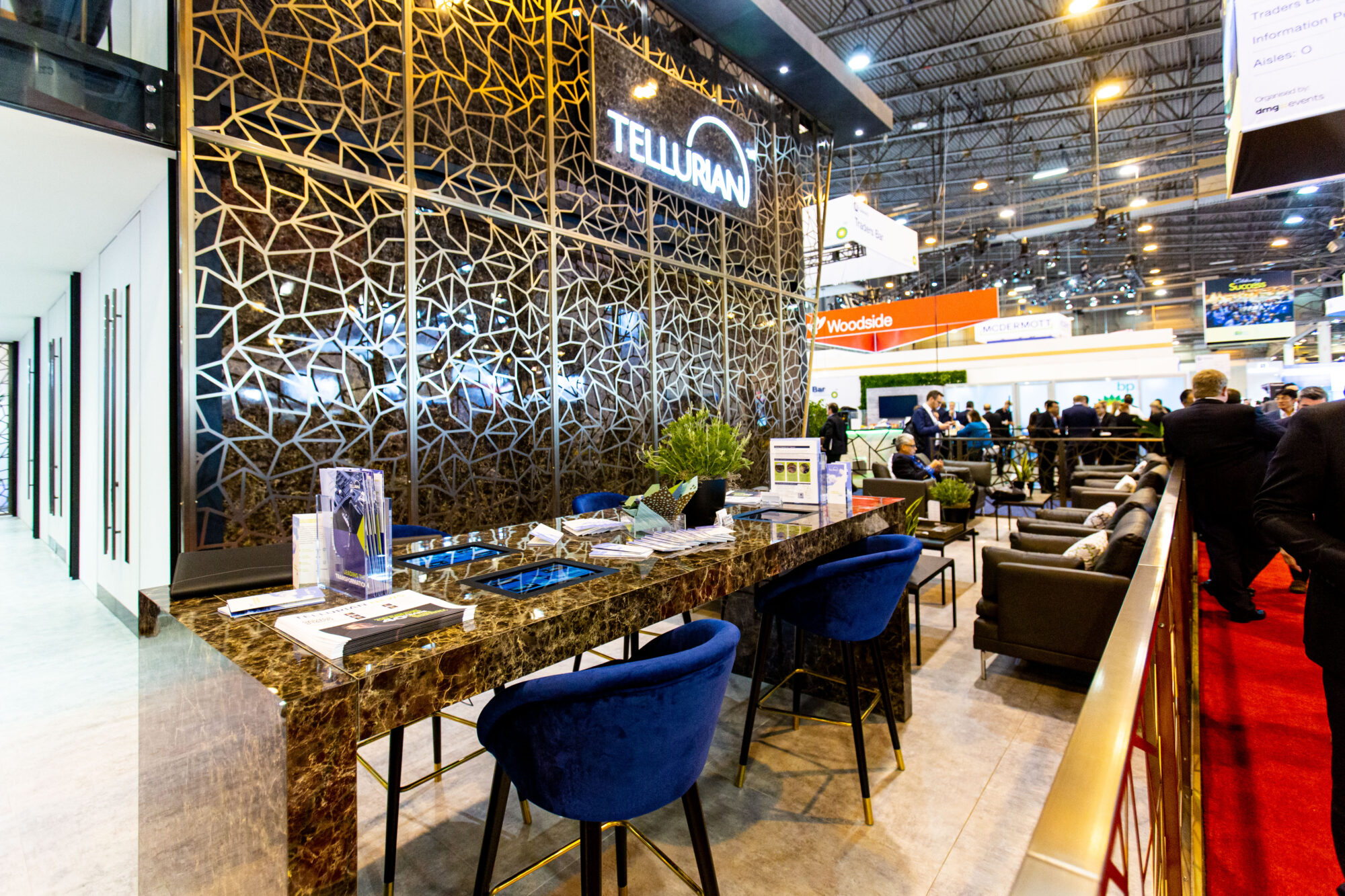 A modern exhibition booth with a large “TELLURAN” sign, decorative geometric wall, marble table with blue velvet chairs, brochures featuring a Tellurian case study, and people networking in a busy, well-lit event hall.