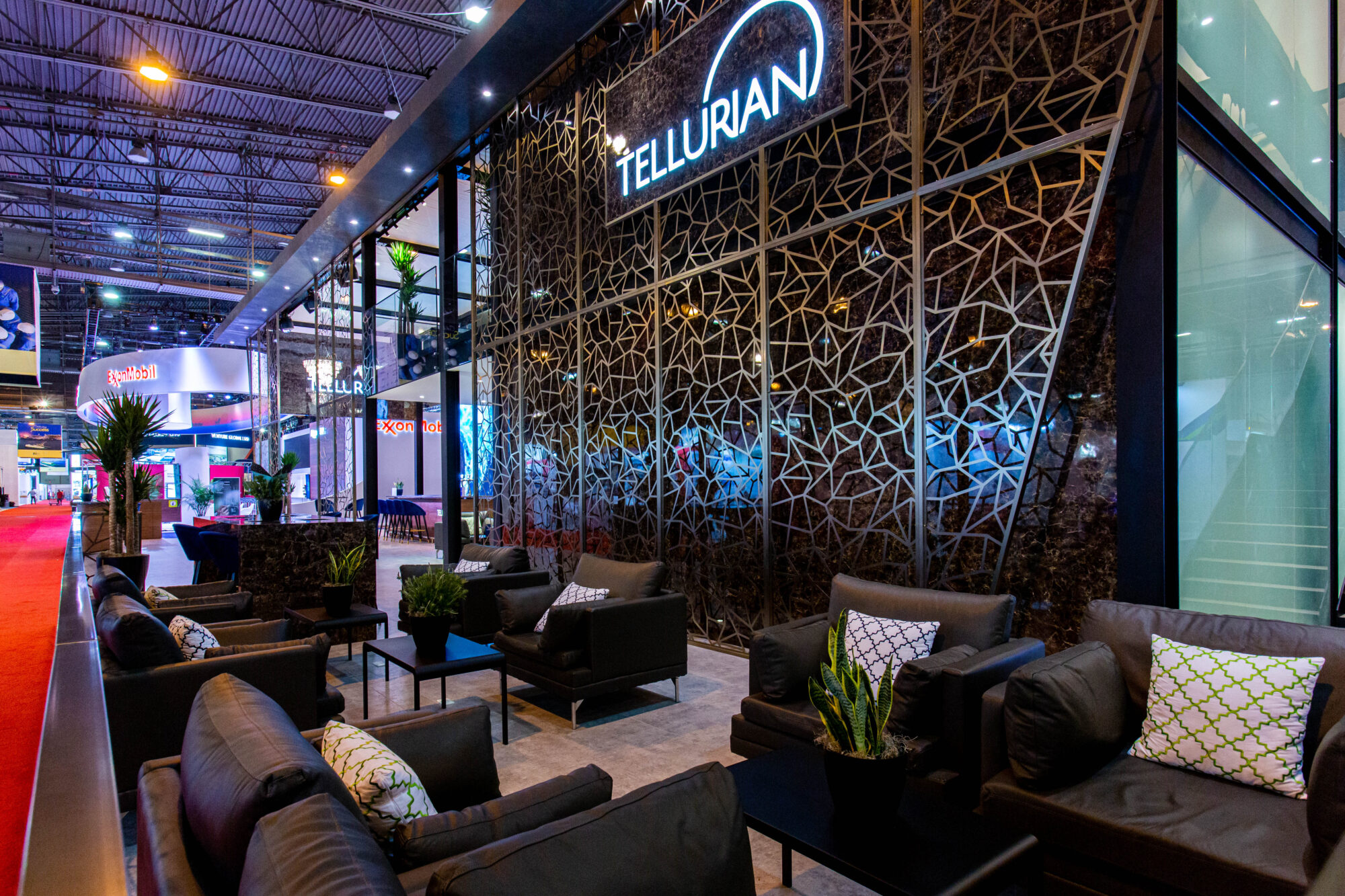 Modern lounge area at an exhibition booth with black leather chairs, green plants, and geometric wall panels. A large illuminated sign reads TELLURIAN, highlighting the space as a showcase for a Tellurian case study. Other booths and lighting are visible in the background.