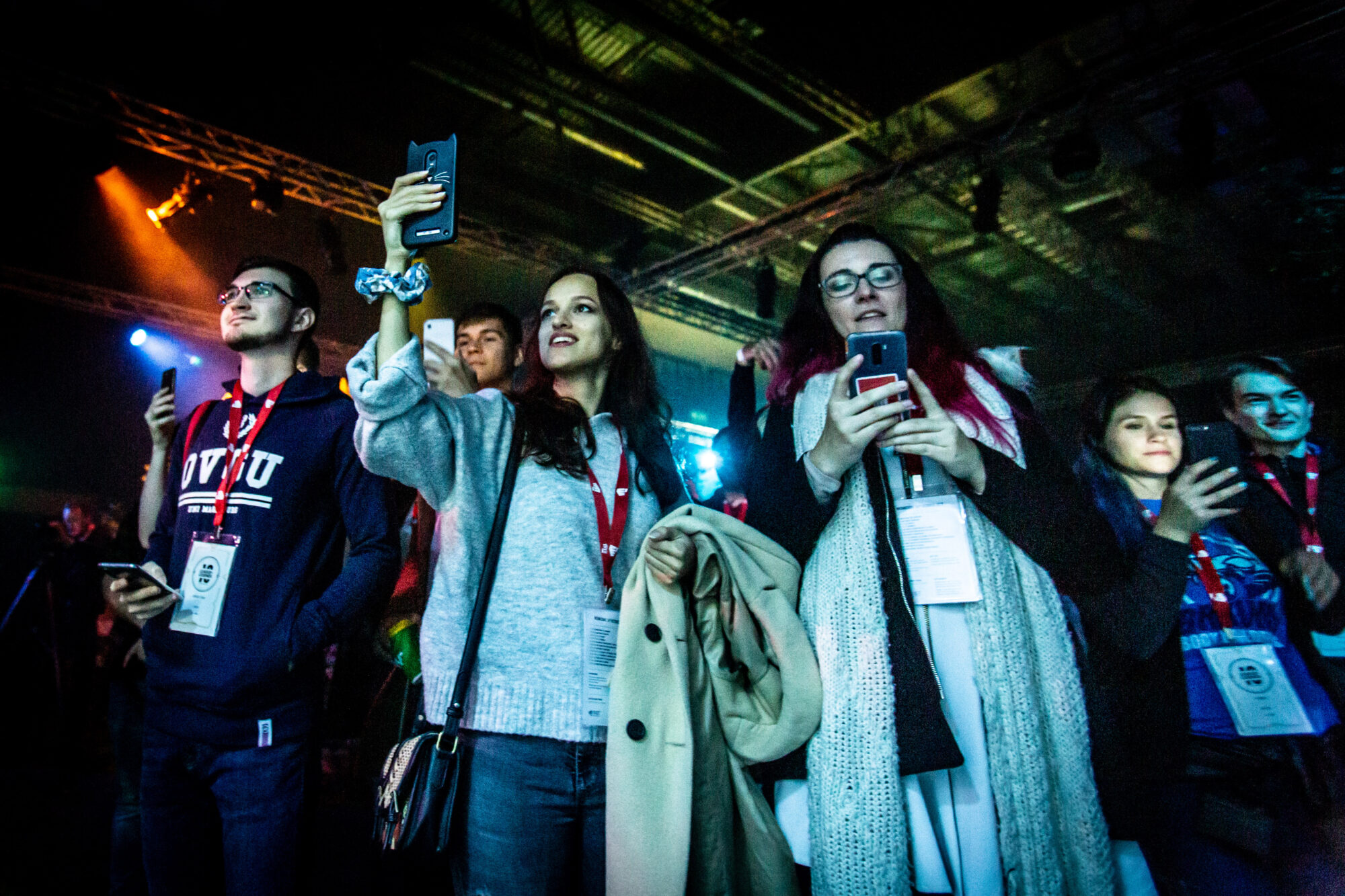A group of young people at a Riot Games L10 indoor event, wearing lanyards and badges, stand under colorful lights. Several are taking photos or videos with their phones while looking toward the stage.
