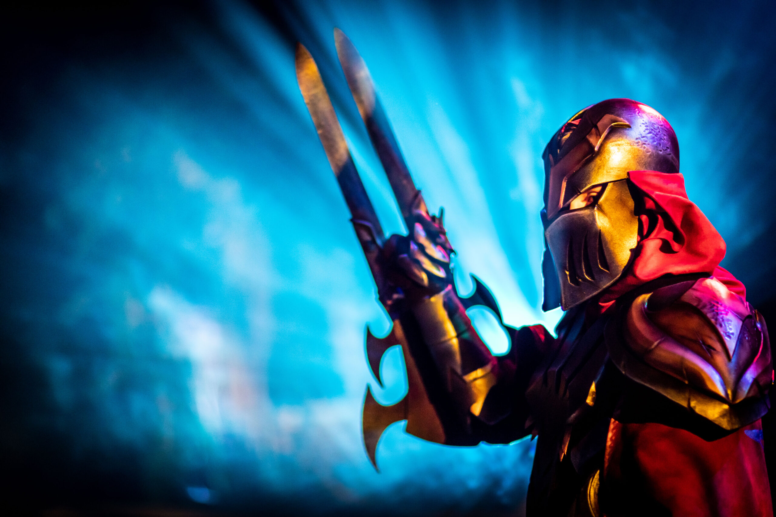 A person in ornate gold and red armor with a horned helmet, inspired by Riot Games L10, holds two large bladed weapons crossed before them. Blue rays of light shine from behind, heightening the dramatic and intense atmosphere.