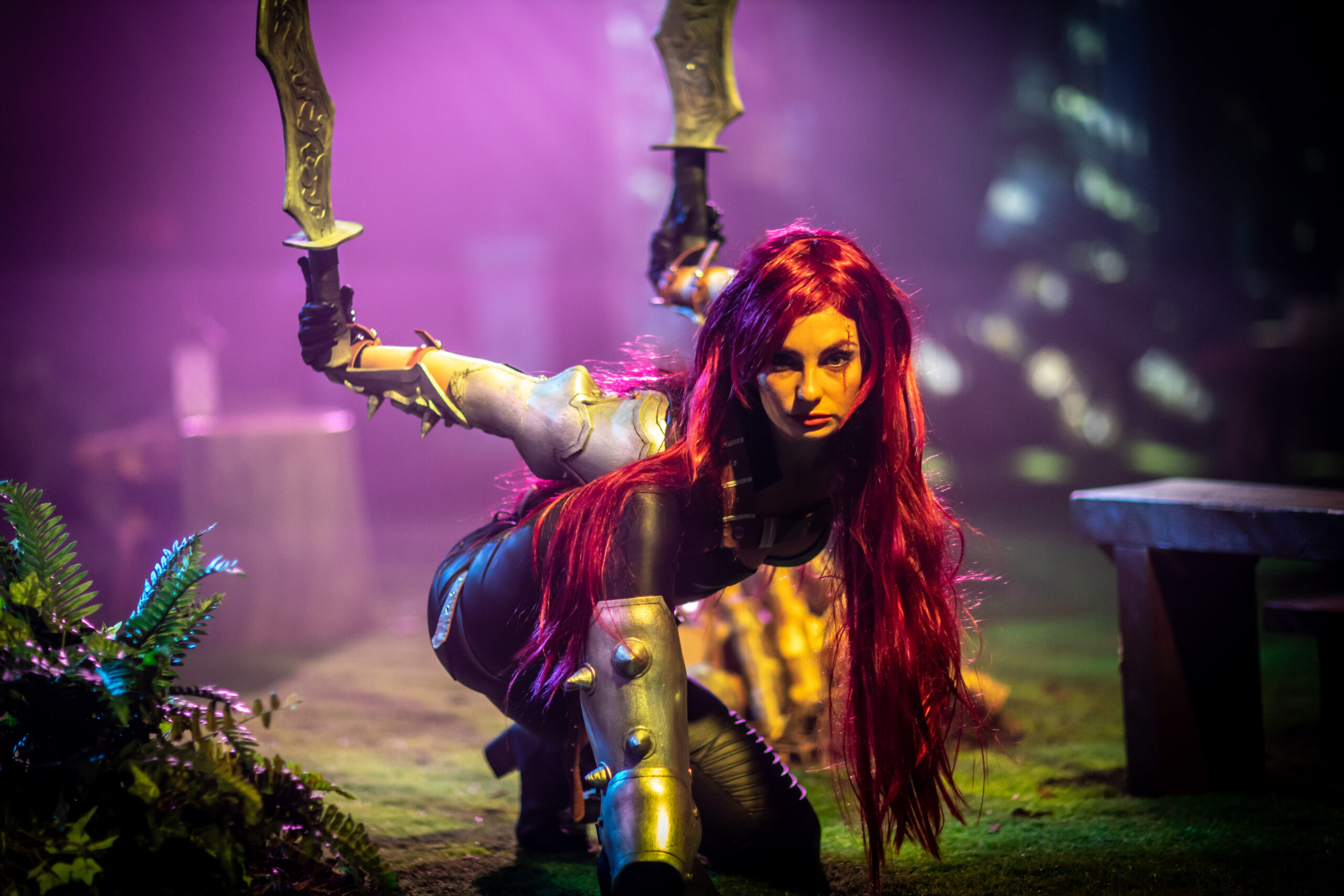 A woman in a fantasy warrior costume with long red hair crouches on the ground, holding two curved swords. Riot Games L10-inspired lighting and greenery surround her, creating an intense, mystical atmosphere.