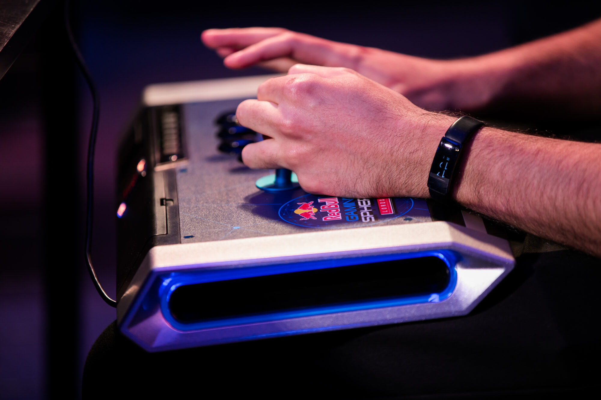Close-up of a person’s hands operating a video game arcade fight stick controller, wearing a fitness tracker on their wrist, with the Red Bull logo and a Red Bull Kumite decal visible on the controller.