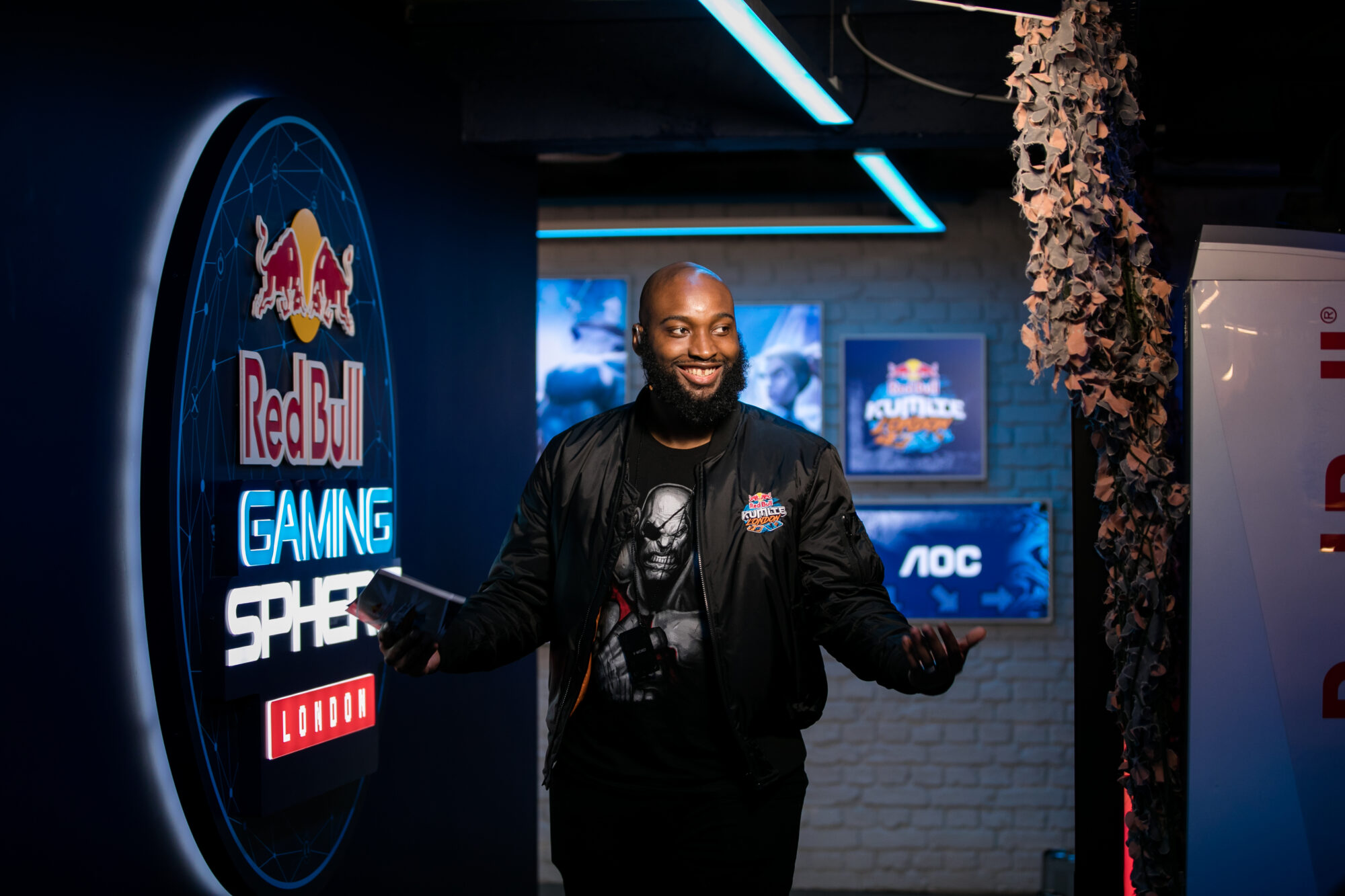 A man in a Red Bull jacket stands smiling with arms outstretched in front of a neon-lit Red Bull Gaming Sphere London sign, blue lighting and gaming posters setting the scene for the energy of Red Bull Kumite.