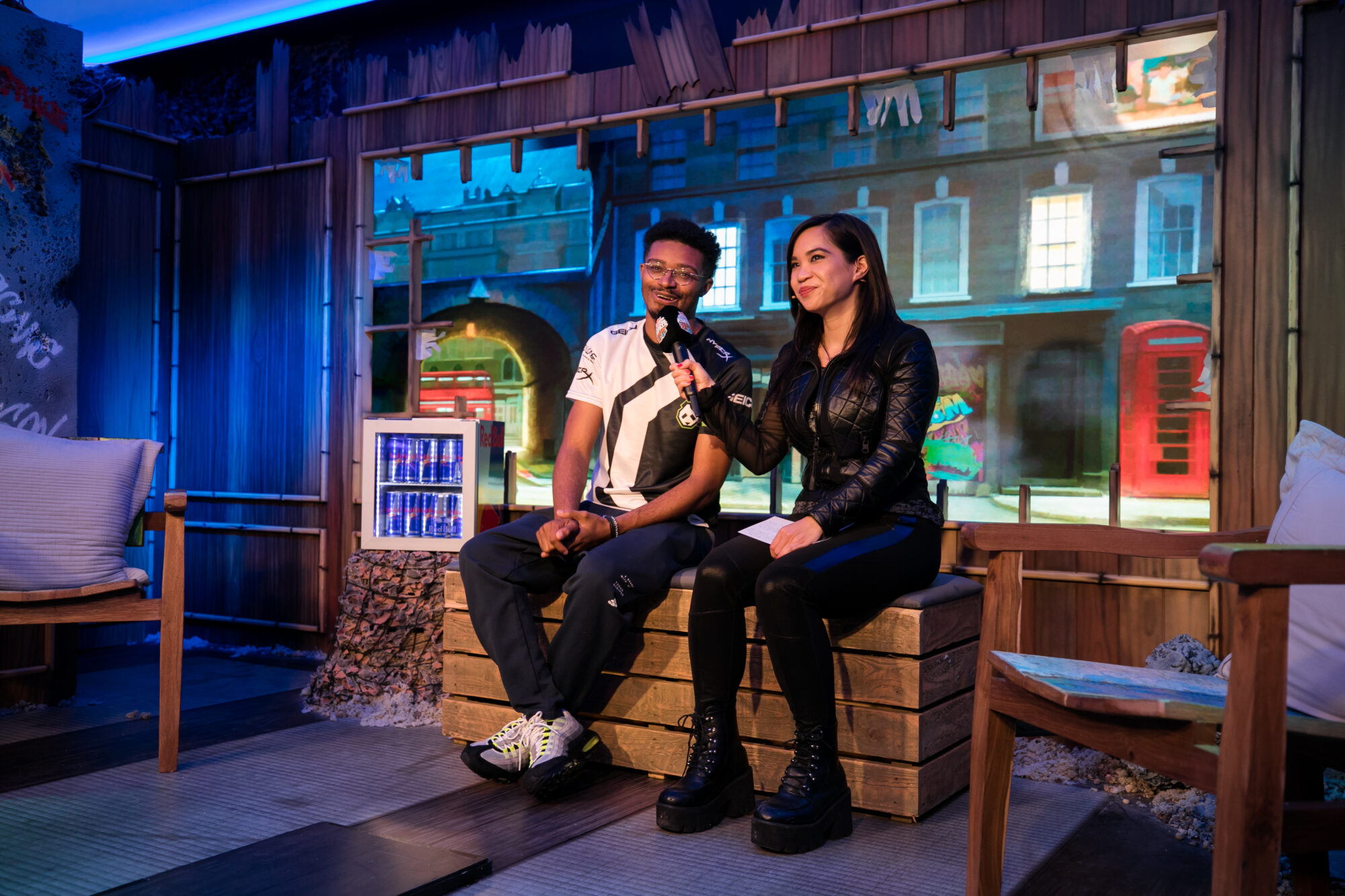 A man and a woman sit on a wooden bench indoors, holding microphones and smiling. The background features a colorful mural of buildings, a red phone booth, and nods to Red Bull Kumite. The setting appears casual and relaxed, with soft lighting.