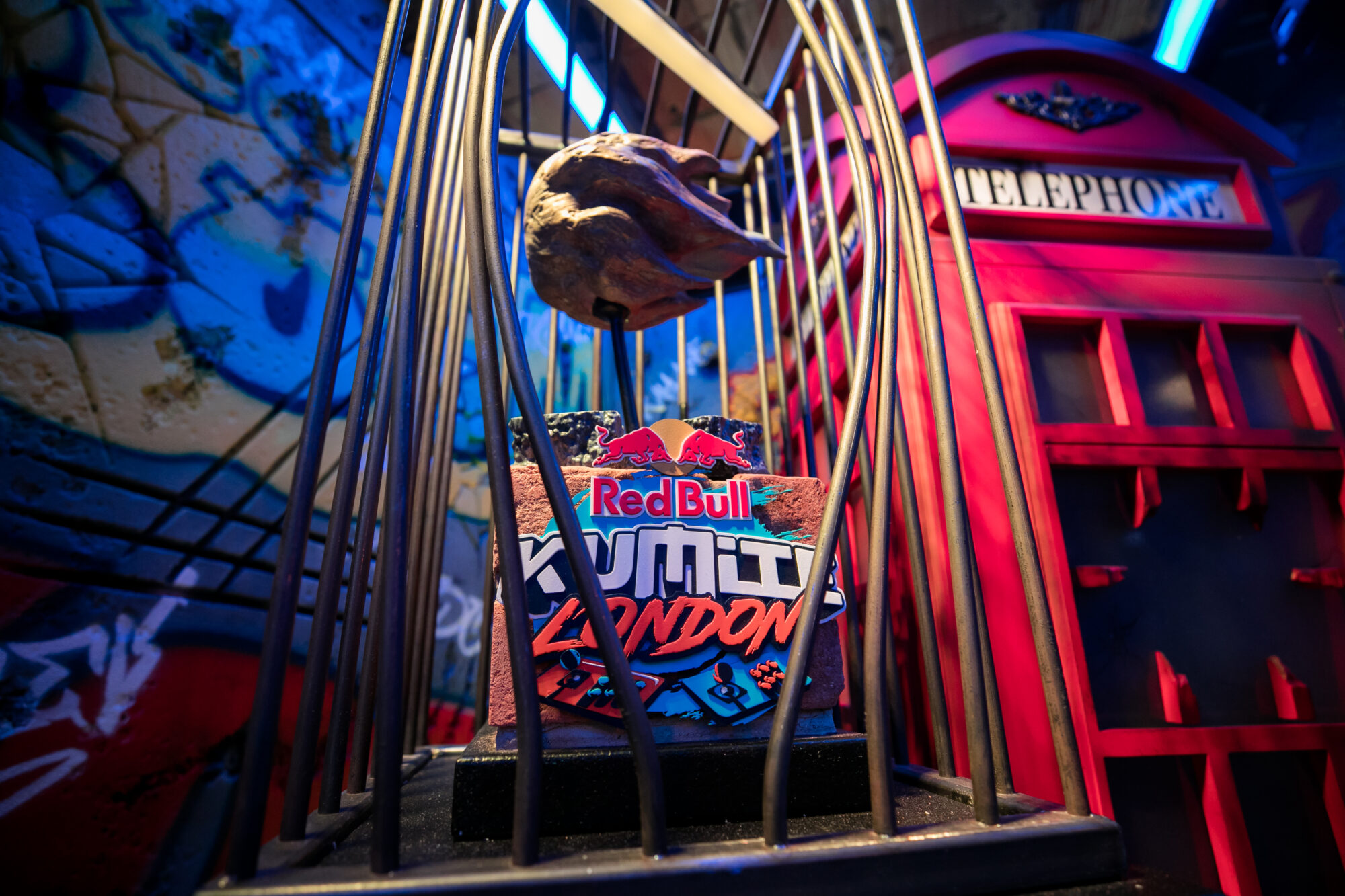 A close-up of a Red Bull Kumite trophy inside a metal cage, with a red British telephone booth and colorful graffiti in the background, all illuminated by vibrant blue and pink lights.