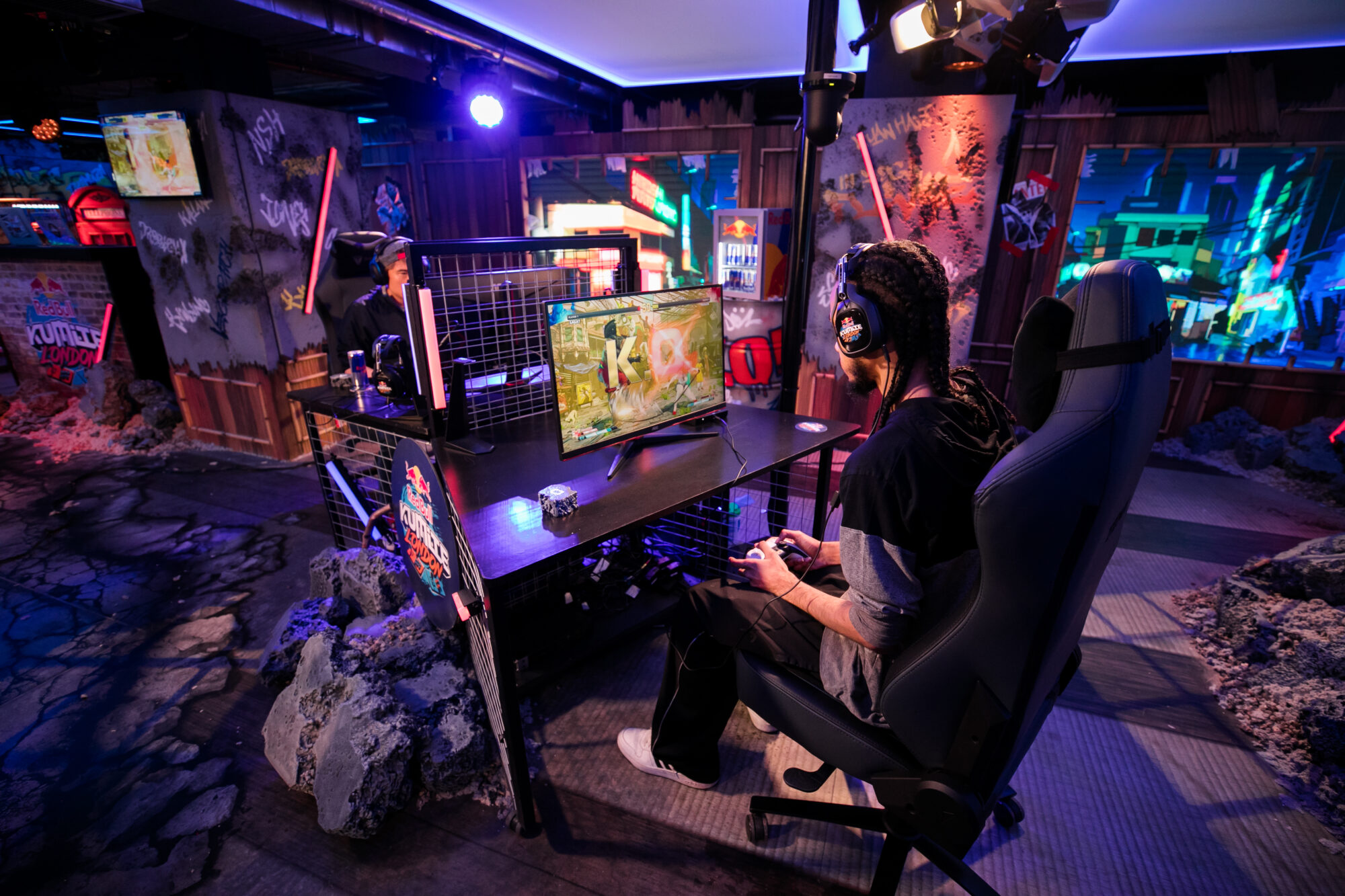 Two gamers sit at separate desks facing monitors, playing a fighting video game. One wears headphones and holds a controller, while the other is partially visible. The rooms colorful lighting and decor evoke a Red Bull Kumite-style energy.
