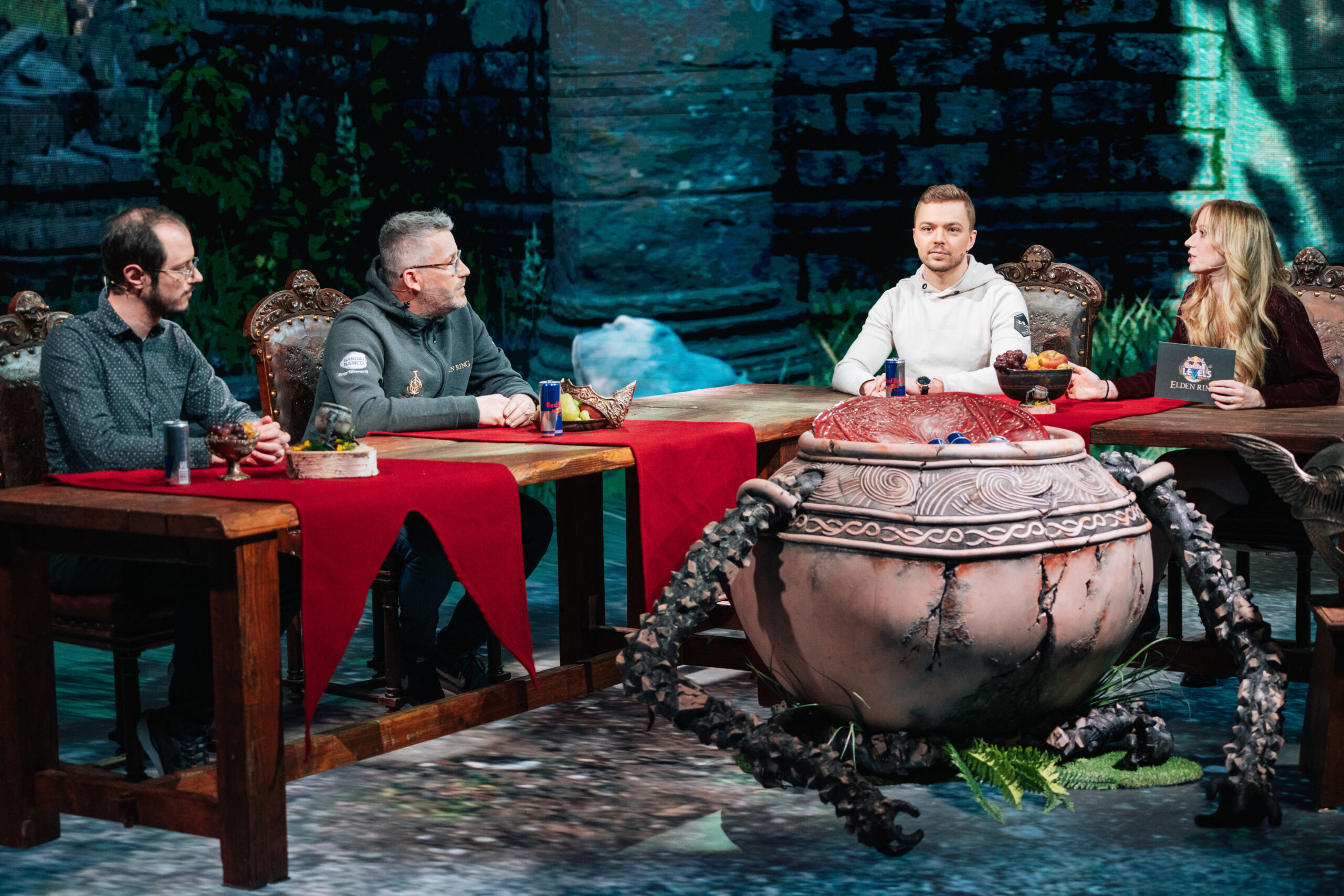 Four people sit around wooden tables with red tablecloths, talking, while a large, ornate, spider-like pot—reminiscent of Elden Ring—stands in the center. Stone walls and greenery in the background create a vivid fantasy setting.
