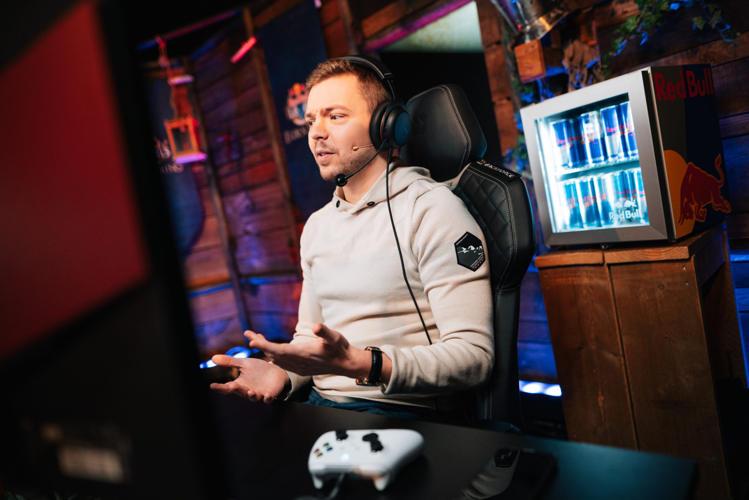 A man wearing headphones and a hoodie sits at a desk with a gaming controller, speaking and gesturing at a monitor—possibly streaming an Elden Ring global XR session. A mini fridge with Red Bull cans is in the background.