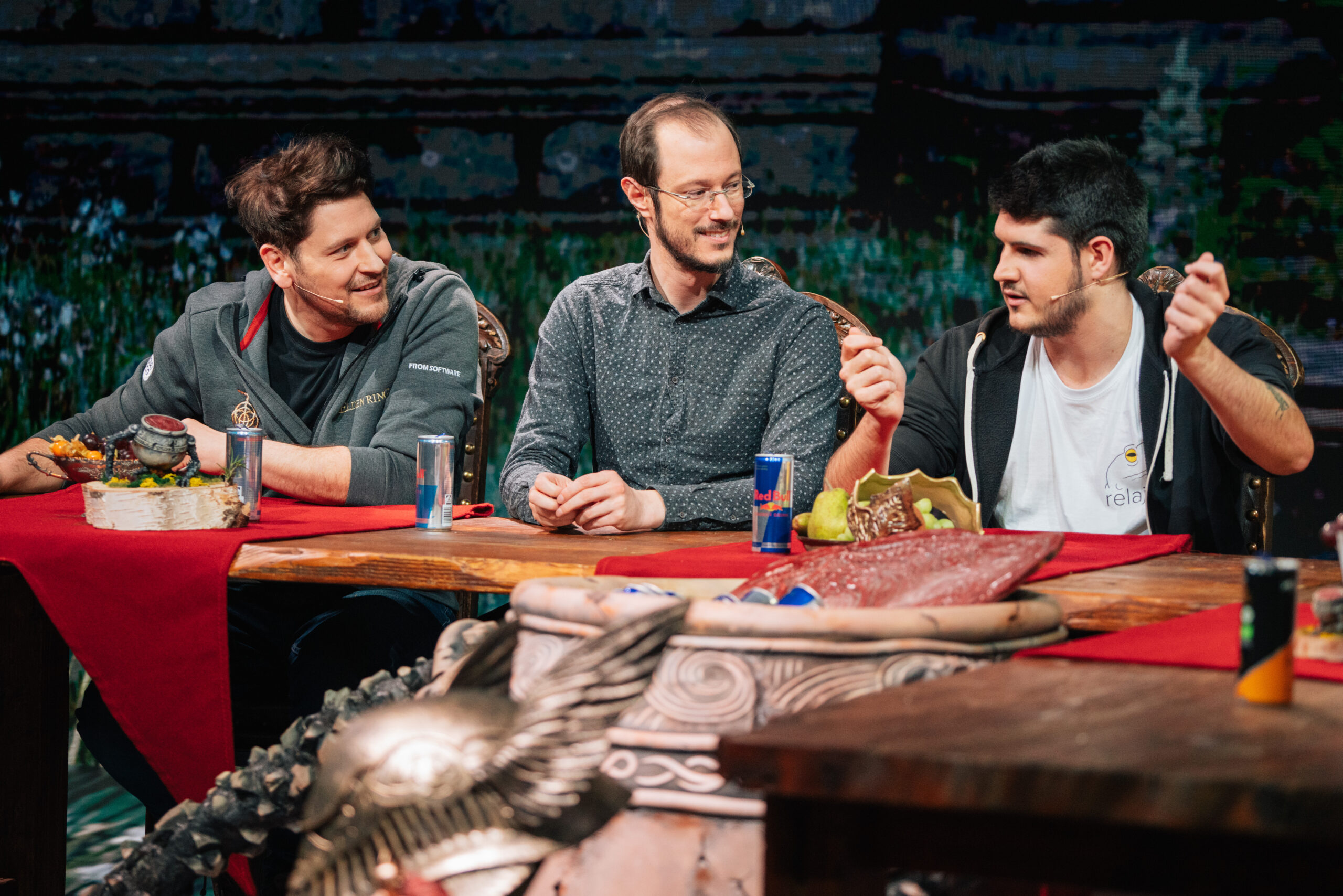 Three men sit at a table covered with red cloth and fantasy-themed props, chatting and smiling. Two cans of Red Bull are on the table. The colorful, blurred background hints at an Elden Ring-inspired global XR fantasy setting.