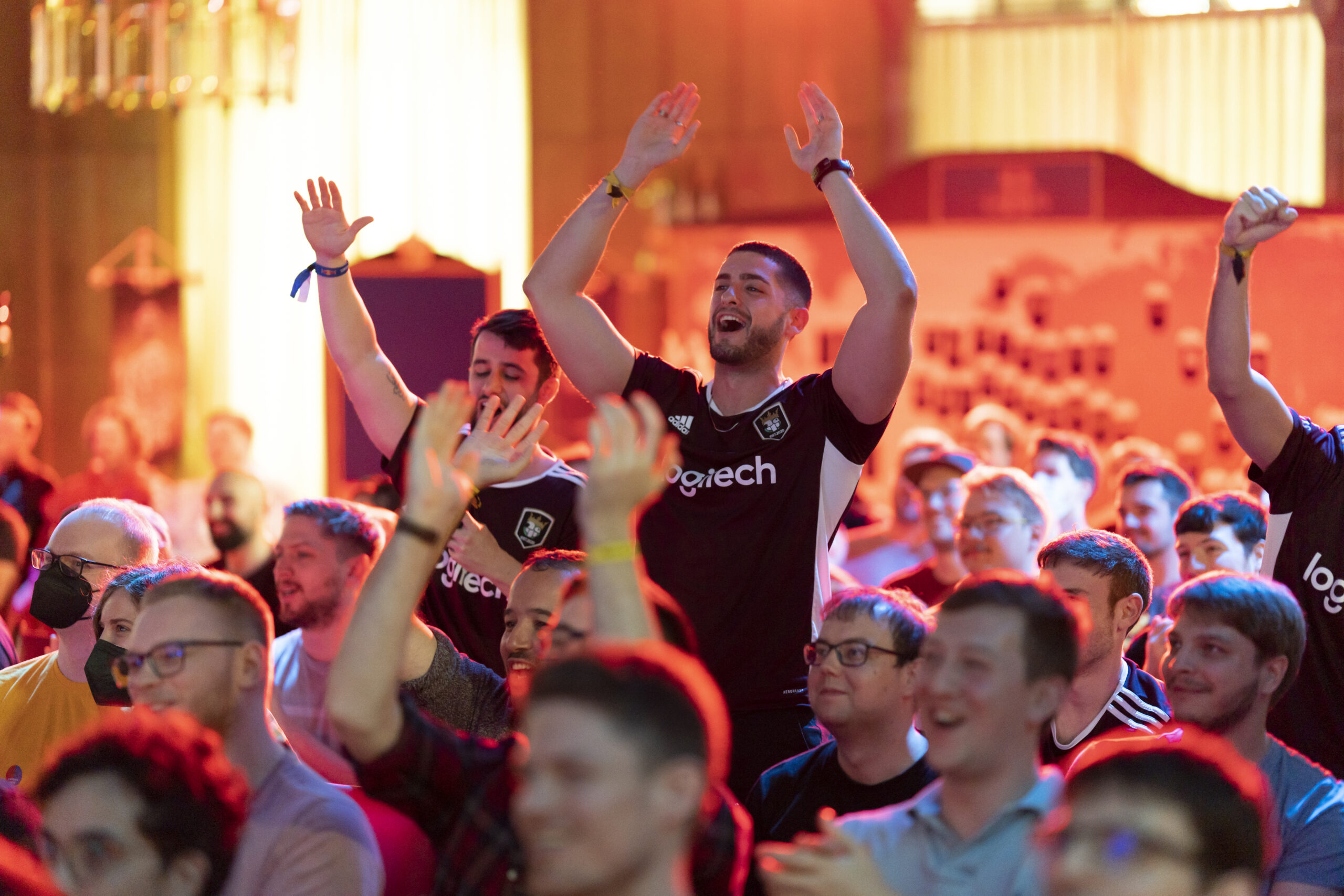 A crowd of enthusiastic fans, many in black Logitech jerseys, cheer with raised hands at an indoor Red Bull Wololo event. The lively atmosphere is filled with smiles and celebration under vibrant, colorful lighting.