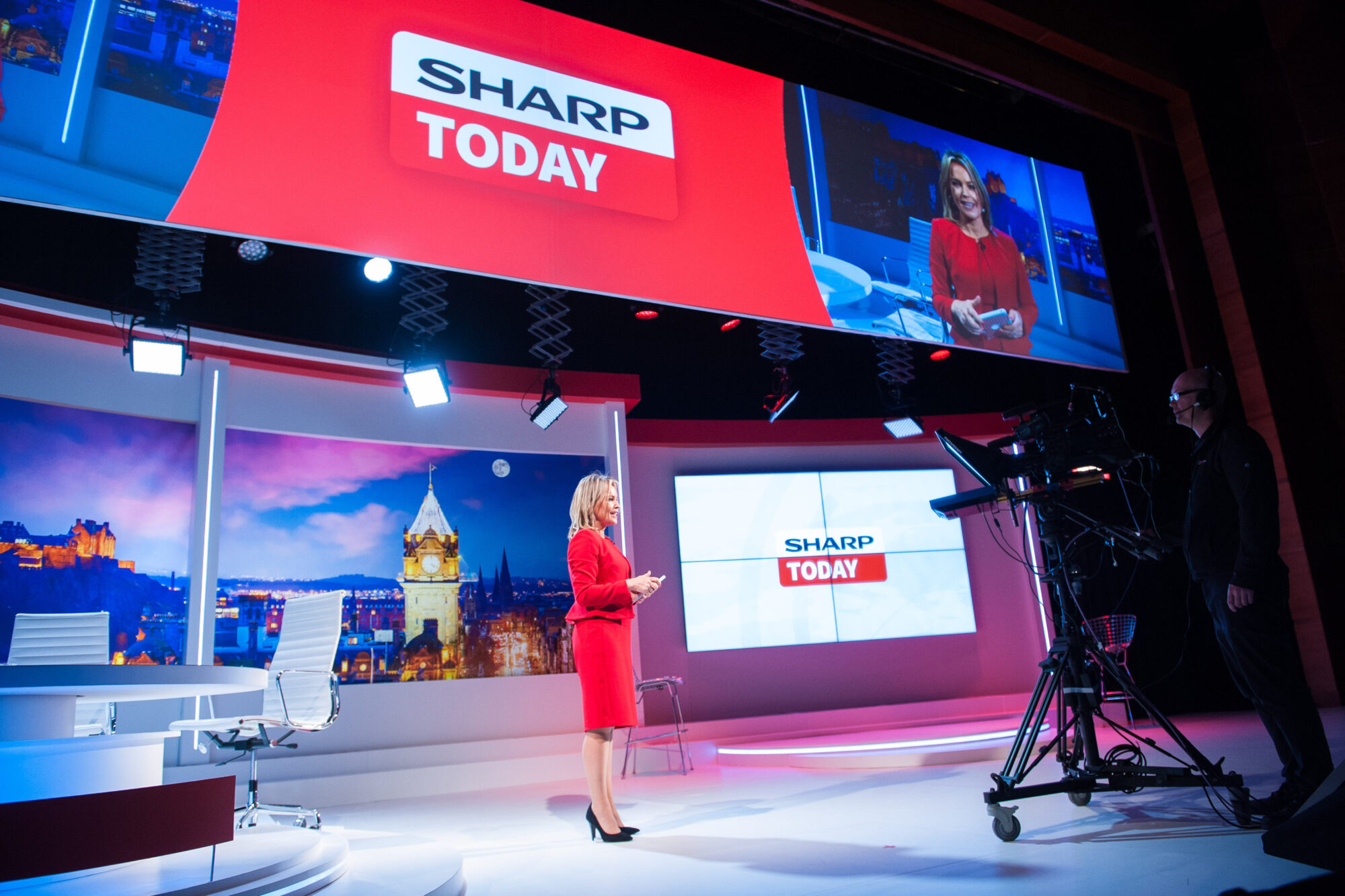 A woman in a red dress stands in a brightly lit TV news studio with “SHARP TODAY” and Inspire Expo displayed on large screens behind her as a cameraman films the scene.