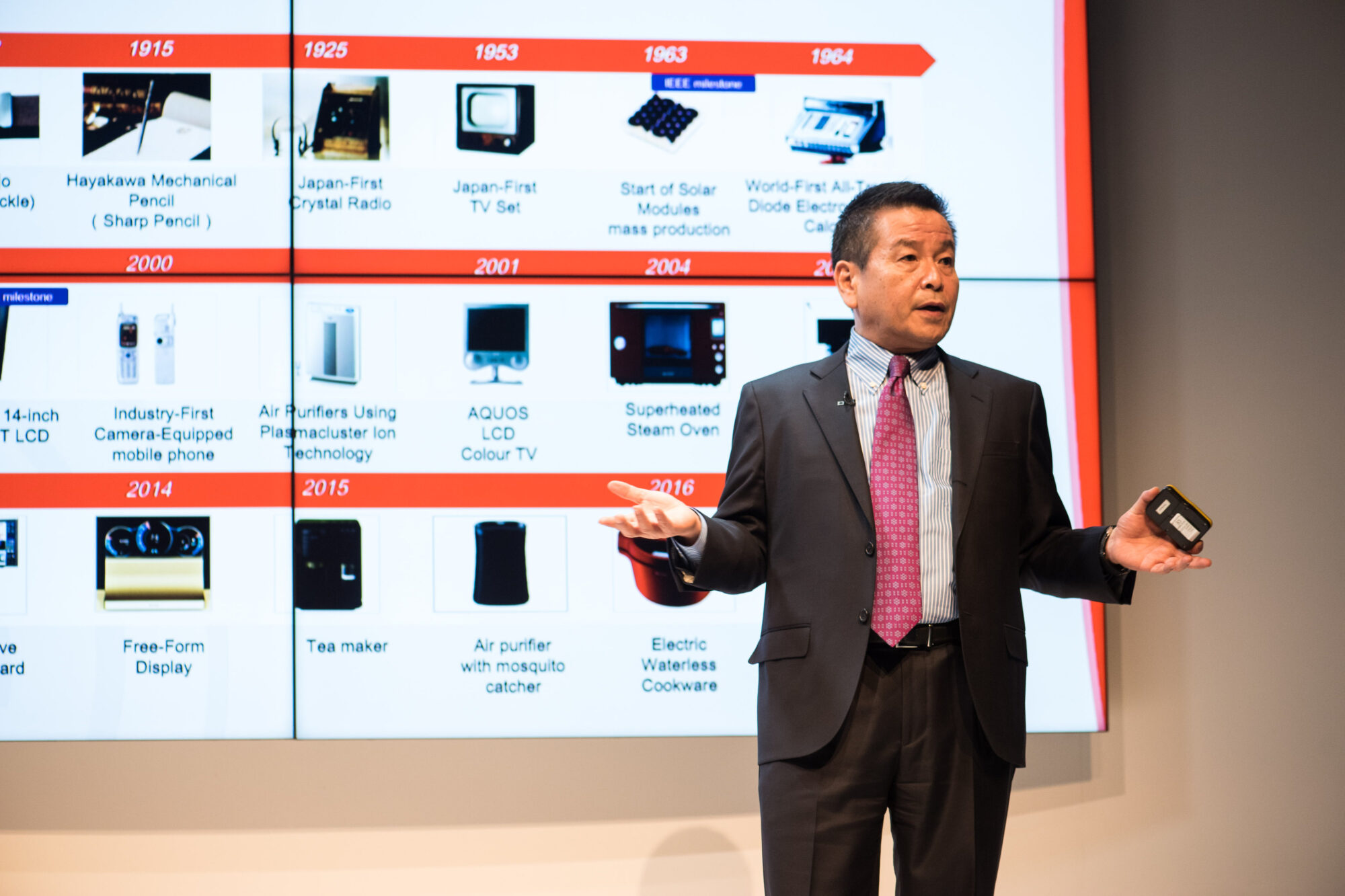 A man in a suit holds a small device while speaking on stage at Inspire Expo, in front of a large timeline slide showcasing technological innovations from 1915 to 2016.