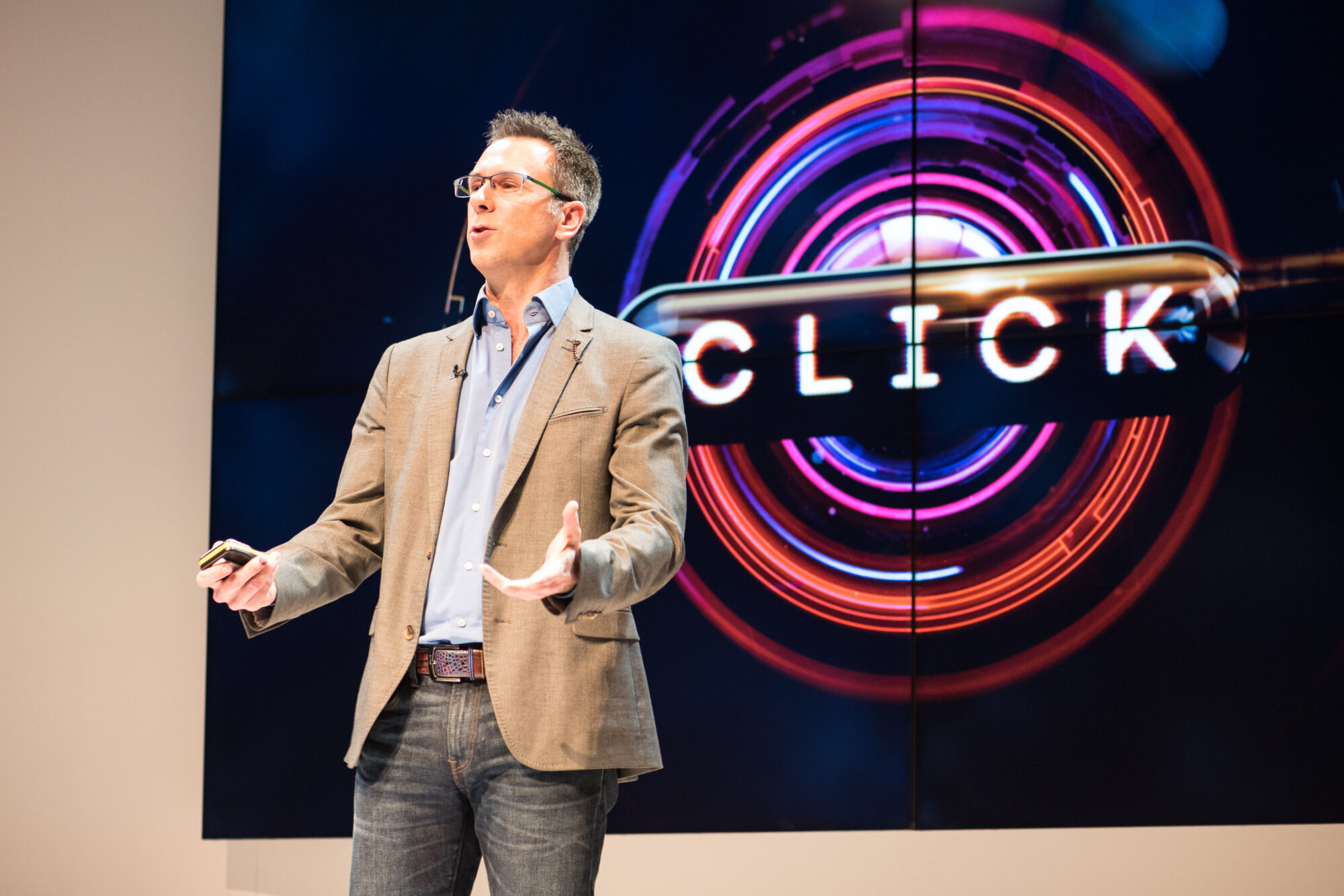 A man in a gray blazer and jeans is speaking on stage at Inspire Expo, gesturing with one hand and holding a device in the other. Behind him, a large screen displays the word CLICK with colorful circular graphics.