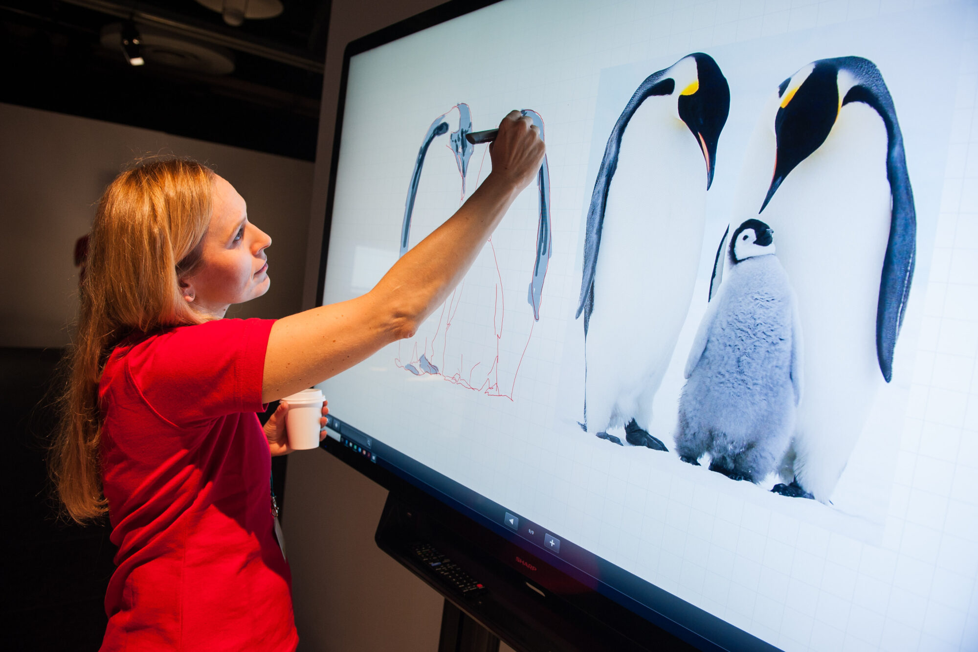 At Inspire Expo, a woman in a red shirt draws an outline of emperor penguins on a large touchscreen, beside a photo of two adult emperor penguins with a chick.
