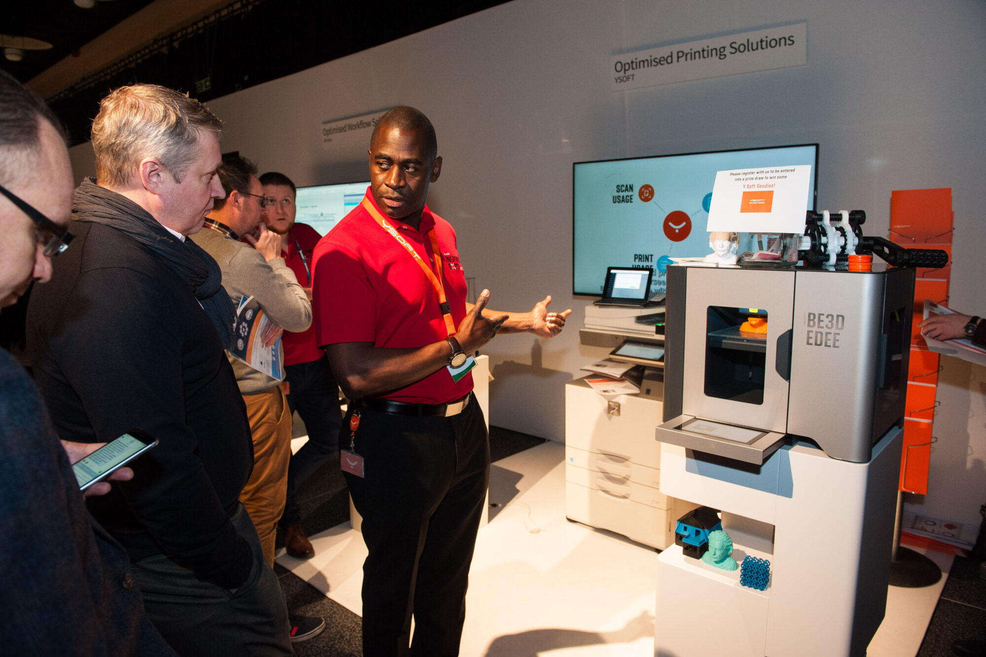 A man in a red shirt explains a 3D printer to a group of people at Inspire Expo, with screens and printed models displayed around the machine.