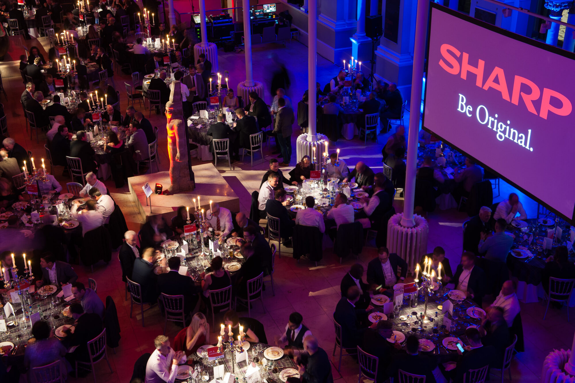 A large, elegant banquet hall with many round tables of people dining by candlelight. At Inspire Expo, a big screen displays the Sharp logo and slogan “Be Original,” while purple and pink lighting bathes the room.