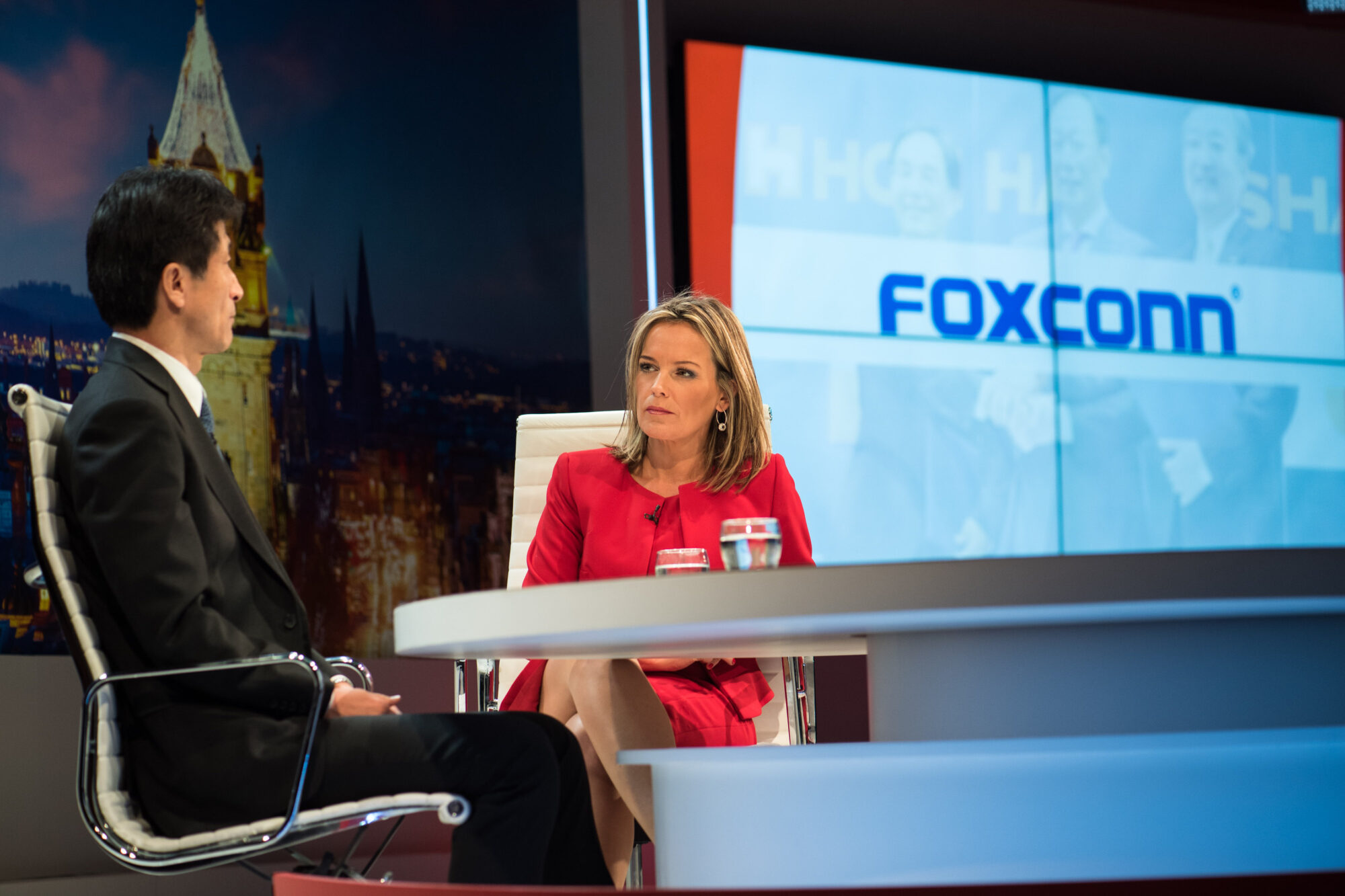A woman in a red blazer interviews a man in a dark suit at a news studio desk, with a large screen behind them displaying the Foxconn logo, blurred images of people, and Inspire Expo highlights.
