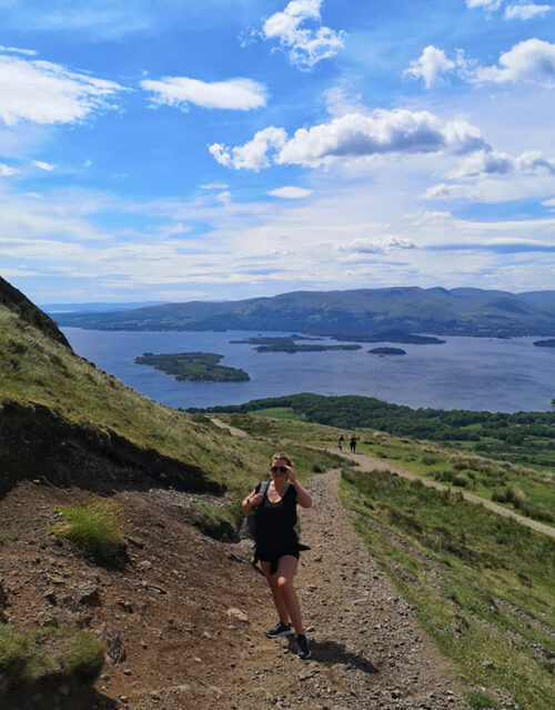 A person in athletic clothes hikes up a rocky trail on a hillside, enjoying a scenic view reminiscent of a marketing team conquering new heights—overlooking a lake, green islands, and distant mountains beneath a blue sky with scattered clouds.