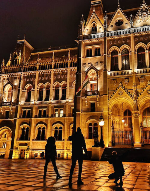 Three silhouetted people pose in front of a grand, ornately lit building at night, featuring arched windows and detailed architecture—an elegant setting perfect for Christmas brand activations, with a flagpole in the center foreground.