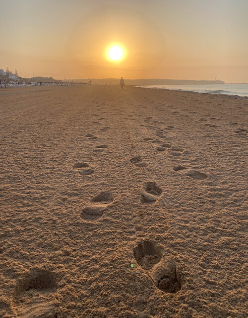 Footprints in the sand lead toward a lone figure—perhaps reflecting like a marketing team brainstorming—walking on a wide, empty beach at sunrise or sunset, with the sun low on the horizon and calm ocean waves to the right.