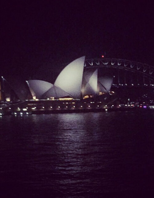 Sydney Opera House illuminated at night, with the Sydney Harbour Bridge in the background, as if awaiting a creative marketing team to capture its shimmering reflections on the dark water in the foreground.