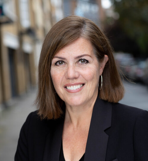 A woman with straight brown hair and hoop earrings smiles at the camera, wearing a black blazer and standing on a city street—an inspiring member of the marketing team, with blurred buildings and cars in the background.