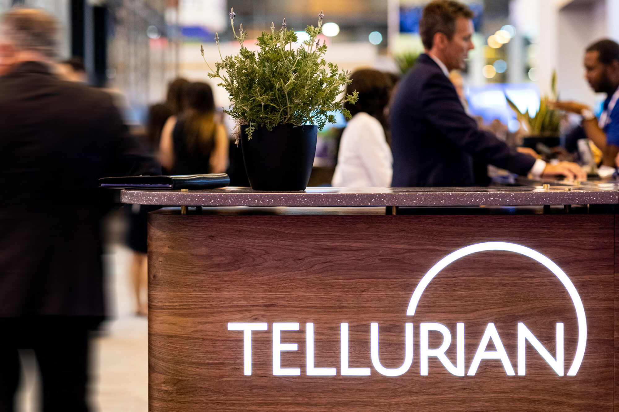 A potted plant sits on a wooden reception desk with the illuminated Tellurian logo, setting the scene for a bustling event where professionals connect—an ideal Tellurian case study in welcoming workspace design.