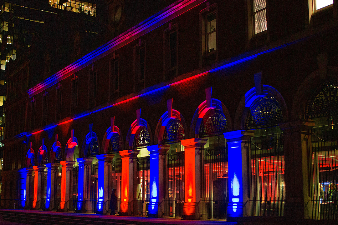 A brick building with large arched windows is illuminated at night with red and blue lights, creating a vibrant, colorful effect on its facade—perfect for celebrating Top Employer Awards.