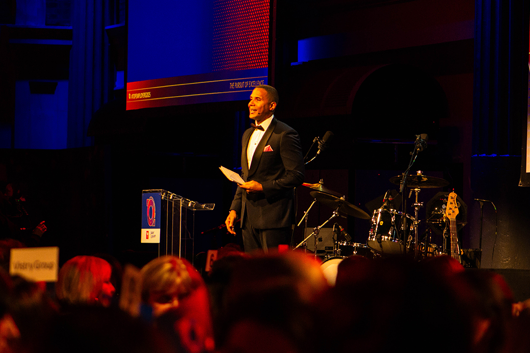 A man in a dark suit stands on stage holding papers, speaking to an audience at the Top Employer Awards. Behind him are drums and musical equipment, with warm lighting illuminating the scene.