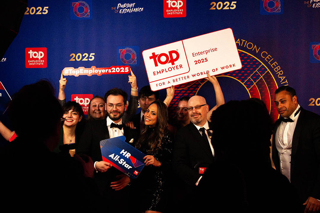 A group of professionally dressed people at an award event pose smiling with signs reading “Top Employer 2025,” “#TopEmployers2025,” and “HR All-Star” against a branded backdrop, celebrating their success at the Top Employer Awards.