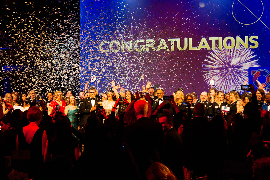 A large group of people in formal attire celebrate on stage as confetti falls, with CONGRATULATIONS and fireworks displayed on a blue and purple screen in the background at the Top Employer Awards.